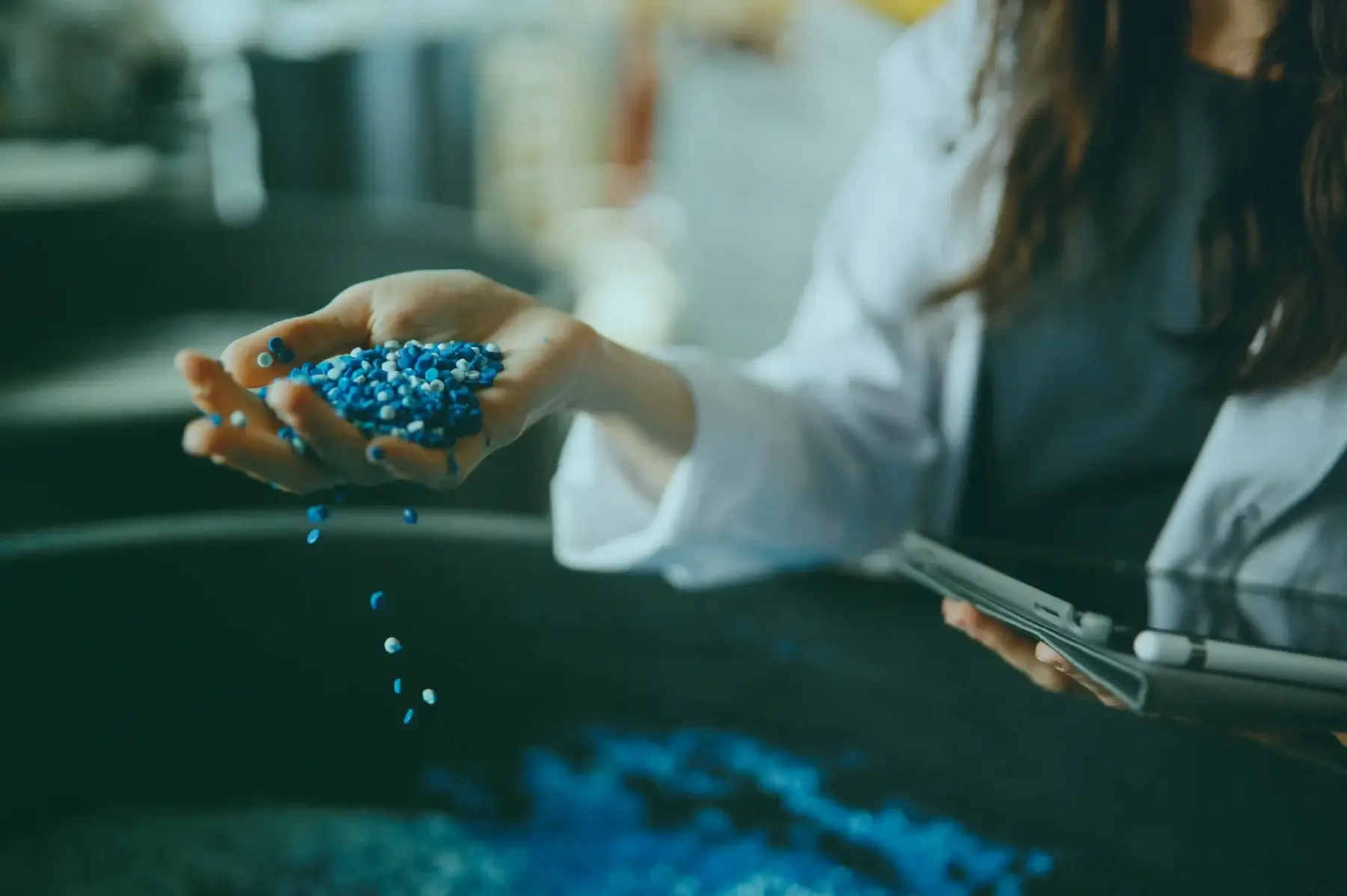 Engineer examining blue plastic pellets while reviewing data on a tablet, representing quality control and sustainable regulatory compliance in the plastics industry.